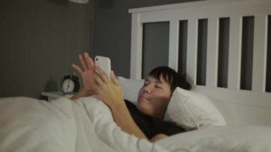 Genç Asyalı adam akıllı telefon kullanıyor. Gece geç saatte yatağında uzanıyor..