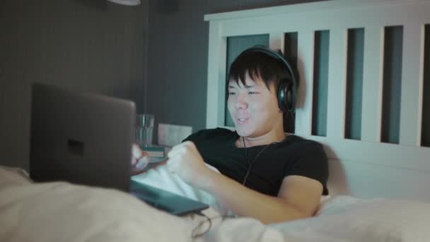 Joven asiático hombre en auriculares usando un portátil tarde en la noche en la cama — Vídeos de Stock