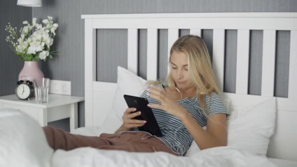 Joven mujer aburrida navegando en las redes sociales en una tableta acostada en una cama en casa — Vídeo de stock