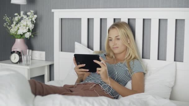 Mujer joven viendo los medios de comunicación en una tableta acostada en una cama en casa — Vídeos de Stock