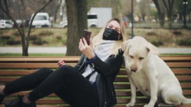 Siyah maskeli genç bir kadın Retriever Labrador köpeğiyle bankta oturur ve parkta selfie çeker. Karantina Coronavirus COVID-19 salgını sırasında şehir parkında.