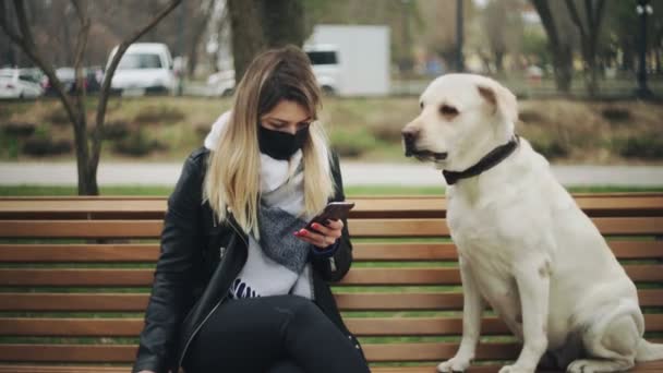 Mujer joven con máscara negra se sienta con perro labrador retriever en el banco y utiliza el teléfono en el parque de la ciudad durante la pandemia coronavirus cuarentena COVID-19 — Vídeos de Stock