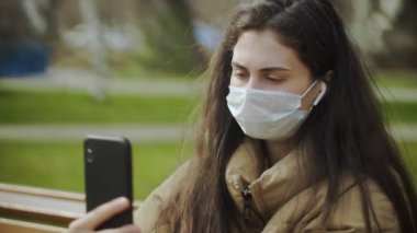 Şehir parkında video kaydı yapmak için akıllı telefon kullanan koruyucu maskeli genç bir kadın. Salgın Covid-19 koronavirüs koruması.
