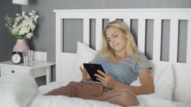 Joven mujer aburrida navegando en las redes sociales en una tableta acostada en una cama en casa — Vídeos de Stock