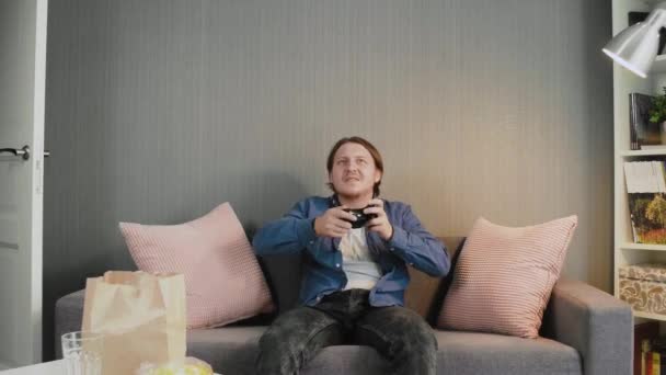 Młody gracz trzymający joysticka i grający w gry wideo siedzi sam na kanapie w domu — Wideo stockowe