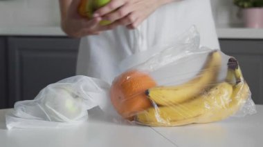 Genç kadın, meyveleri plastik torbadan yeniden kullanılabilir ağ torbasına, mutfak ahşap masasına taşıyor. Sıfır atık ev ve çevre torbası. Çevre Alışverişi.