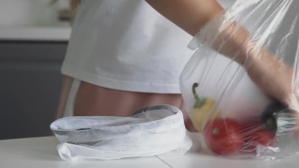 若い女性の閉じるキッチン木製のテーブルの上に再利用可能なメッシュバッグにビニール袋から野菜を移動します。ゴミゼロの家とエコバッグ。エコショッピング. — ストック動画