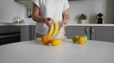 Genç bir kadın, portakalları şeffaf bir çevre çantasına muz elması koyar. Sıfır atık. Plastik hayat yok. Sağlıklı fitness yaşam tarzı. Geri dönüşüm kavramı.