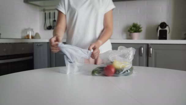 Νεαρή γυναίκα μετακινήστε τα λαχανικά από την πλαστική σακούλα στην επαναχρησιμοποιήσιμη τσάντα ματιών στο ξύλινο τραπέζι της κουζίνας. Μηδέν απόβλητα σπίτι και οικολογική τσάντα. Οικολογικές αγορές. — Αρχείο Βίντεο