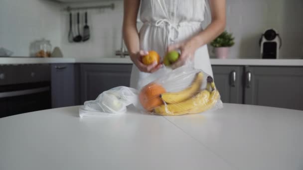 तरुण स्त्री स्वयंपाकघरातील लाकडी टेबलवर पुन्हा वापरण्यायोग्य जाळीच्या पिशवीत प्लास्टिकच्या पिशवीतून फळे हलवते. झिरो कचरा घर आणि इको बॅग. इको खरेदी . — स्टॉक व्हिडिओ