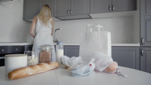 Slide shot av pakket ut handlepose med brød, havremel, bokhvete, melk, grønnsaker og frukt på trebord på kjøkkenet. Kvinne på backgounds. Null sløsing med hjem og miljøveske. Øko-shoppingen . – stockvideo