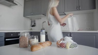 Sarışın kadın ekmek, yulaf ezmesi, süt ve sebzeleri masadan rafa taşıyor. Sıfır atık ev ve çevre torbası. Çevre Alışverişi.
