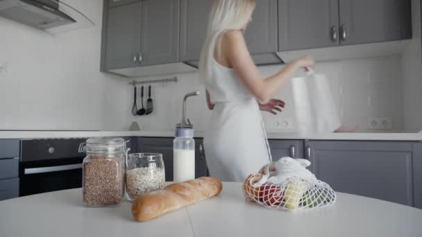 Blond kvinna flyttar bröd, havregryn, mjölk, grönsaker från bord till hyllor. Noll avfall hem och eko väska. Ekologisk handel. — Stockvideo