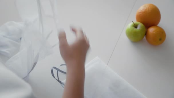 Over de schouder van een vrouw die op houten tafel een boodschappentas met appels, sinaasappels en citroen uitpakt. Geen afval thuis en eco tas. Eco-winkelen. — Stockvideo