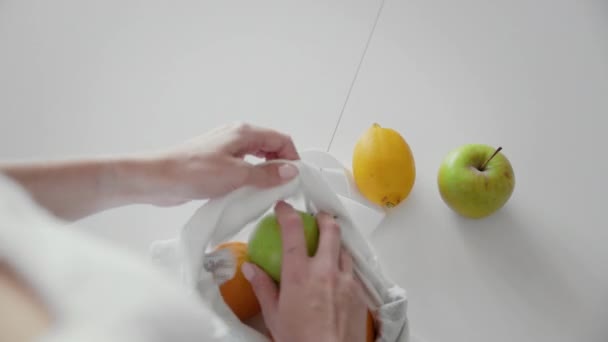 Over de schouder uitzicht van de vrouw verpakking stof boodschappentas met appels, sinaasappels en citroen op de keuken op houten tafel. Geen afval thuis en eco tas. Eco-winkelen. — Stockvideo