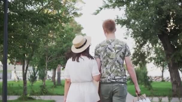 年轻貌美的夫妻手拿着购物袋在夏天的公园里散步 — 图库视频影像