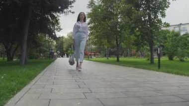 Genç bir kadın elinde sebzelerle pamuk örgüsü alışveriş torbası tutuyor ve yaz parkında yürüyor. Sıfır atık.