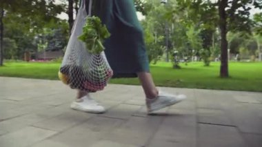 Yeşil etekli ve beyaz spor ayakkabılı genç bir kadının yandan görünüşü sebzeli pamuk örgüsü alışveriş torbası tutuyor ve yaz parkında yürüyor. Sıfır atık.