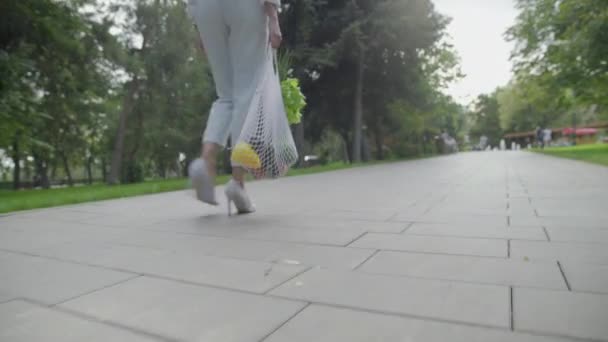 Baksidan av ung kvinna i blå byxor och vita skor håller bomull mesh shoppingväska med grönsaker och promenader i sommarparken. Handeln med nollavfall. — Stockvideo