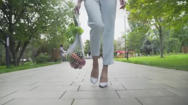 Framsidan av ung kvinna i blå byxor och vita skor håller bomull mesh shoppingväska med grönsaker och promenader i sommarparken. Handeln med nollavfall. — Stockvideo