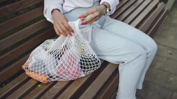 Jonge vrouw zit op de bank en neemt tomaat uit katoenen mesh boodschappentas in het zomerpark. Geen afval winkelen. — Stockvideo