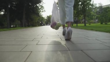 Mavi pantolonlu ve beyaz ayakkabılı genç bir kadının arka manzarasında pamuk örgüsü ile meyve dolu alışveriş torbaları ve yaz parkında yürüyüşler var. Sıfır atık.