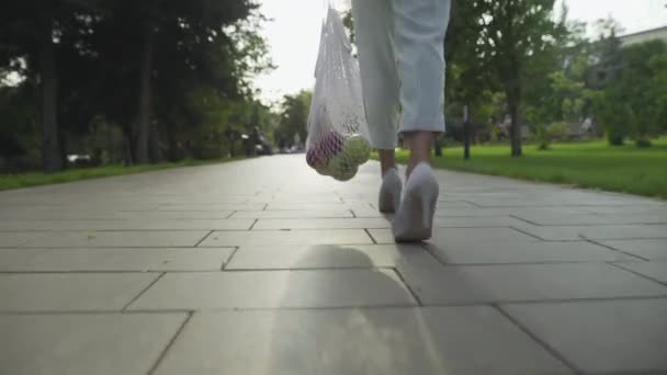 Вид сзади молодой женщины в синих брюках и белых туфлях держит хлопчатобумажную сетку торговый мешок с фруктами и прогулки в летнем парке. Нулевые отходы . — стоковое видео