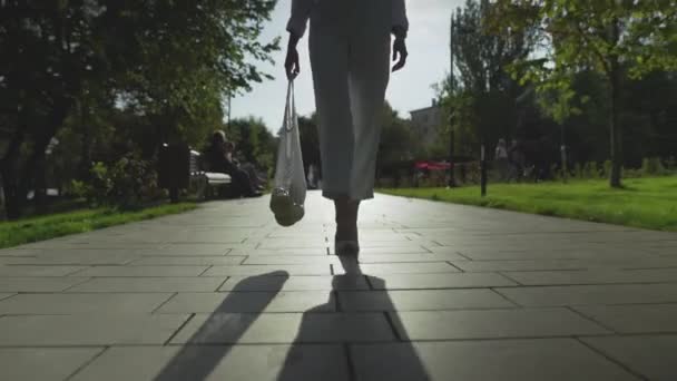 Elölnézetben nő tartja pamut háló bevásárló táska gyümölcsökkel és séták a nyári parkban — Stock videók