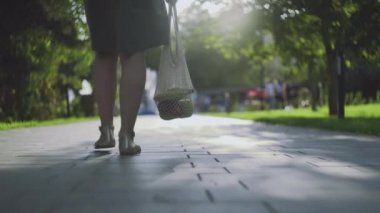 Genç bir kadın elinde sebzelerle pamuk örgüsü alışveriş çantası tutuyor ve yaz parkında yürüyor.