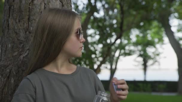 Junges Mädchen mit Sonnenbrille trinkt Orangensaft im Sommerpark — Stockvideo