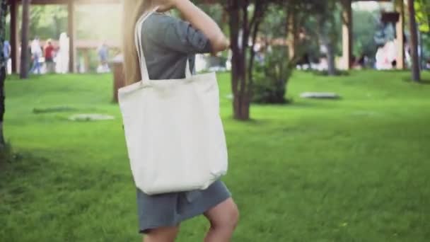 Zijaanzicht van Jong meisje houdt witte textiel eco tas en wandelingen in het zomerpark — Stockvideo