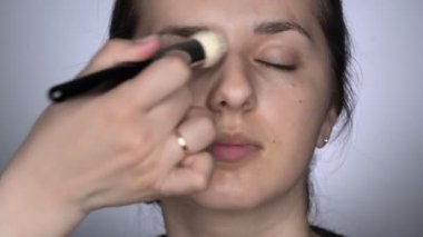 Stüdyoda oturan güzel ve muhteşem kadın için profesyonel makyaj süreci. Makyaj sanatçısı makyaj fırçası kullanarak cilt bakım kremi sürüyor