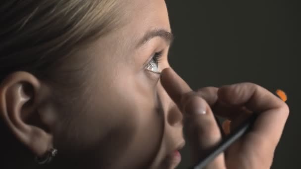 Πλευρική άποψη του make-up artist κάνοντας επαγγελματικό make-up για τη νεαρή γυναίκα στο στούντιο ομορφιάς. Make up Καλλιτέχνης εφαρμόζει concealer στην περιοχή των ματιών — Αρχείο Βίντεο