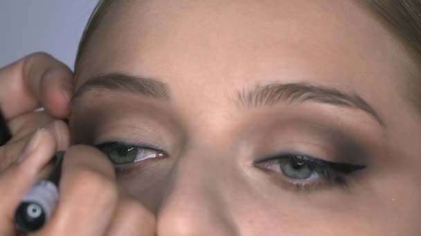 Μακρο πλάνο του καλλιτέχνη μακιγιάζ κάνει επαγγελματική make-up για νεαρή γυναίκα στο στούντιο ομορφιάς. Μακιγιάζ Καλλιτέχνης αντλεί βέλη πάνω από το μάτι, eyeliner — Αρχείο Βίντεο