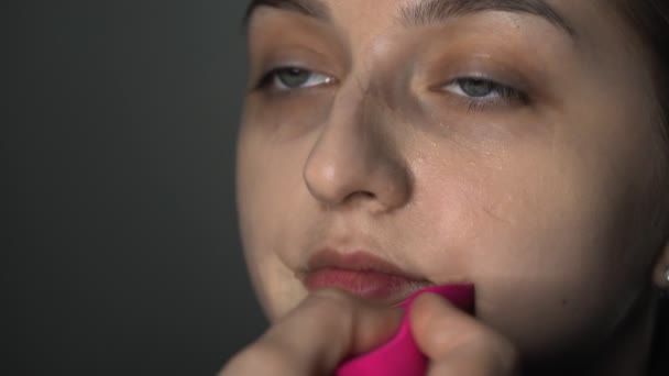 美容室で若い女性のためのプロの化粧を作るメイクアップアーティストのクローズアップ。メイクアーティストは顔にコンシーラーを適用するために特別なスポンジを使用しています — ストック動画