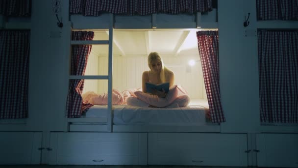 Женщина в ночном белье читает книгу перед сном в уютной комнате общежития — стоковое видео