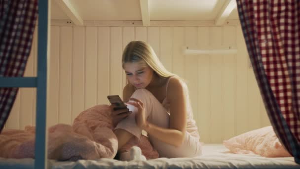 Close-up van jonge aantrekkelijke vrouw zitten op het bed in het hostel kamer en met behulp van smartphone 's nachts — Stockvideo