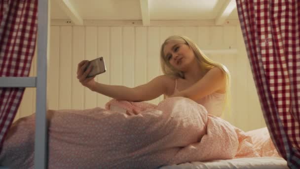 Gros plan de la jeune femme attrayante assise sur le lit dans la salle de l'auberge et en utilisant smartphone ayant chat vidéo — Video