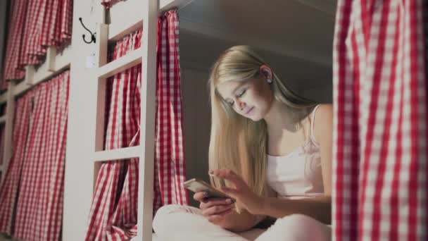 Mujer atractiva joven sentado en la cama en la habitación del albergue y el uso de teléfono inteligente — Vídeos de Stock