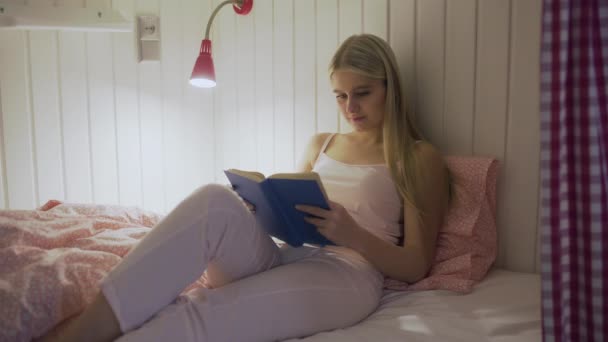 Una joven mujer bonita está leyendo un libro mientras está acostada en una cama por la noche — Vídeo de stock