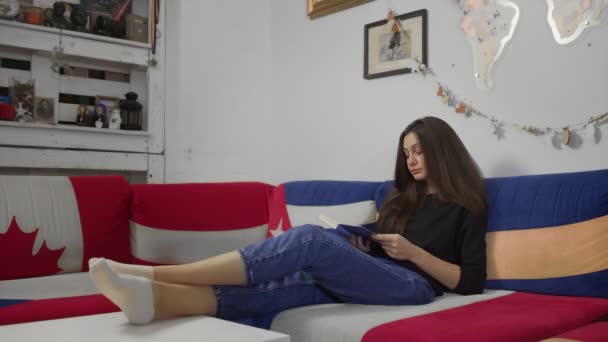 Giovane donna che legge libro di carta seduta in un accogliente divano a casa — Video Stock