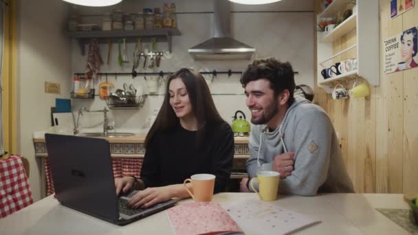 Pareja sonriente tomando café en la cocina. Pareja alegre viendo la computadora portátil. Hombre feliz hablando con la mujer en la cocina. — Vídeos de Stock