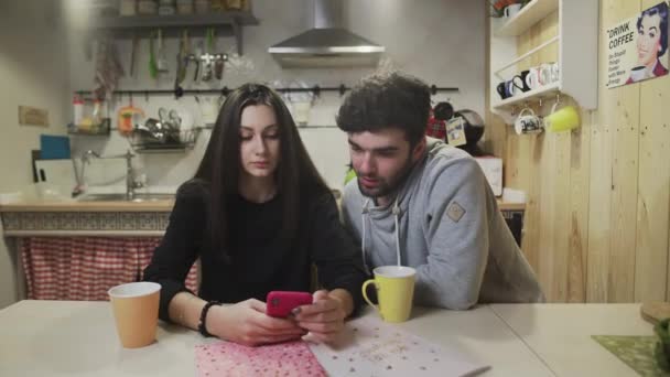 젊은 부부가 부엌에서 아침을 먹고 있습니다. 스마트폰 화면을 보는 즐거운 커플. 행복 한 남자가 부엌에서 여자와 이야기하는 모습. — 비디오