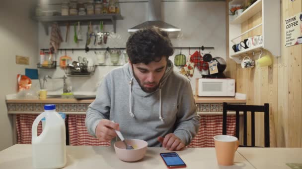 Молодий чоловік снідає на кухні. Людина їсть кукурудзяні пластівці і користується смартфоном — стокове відео