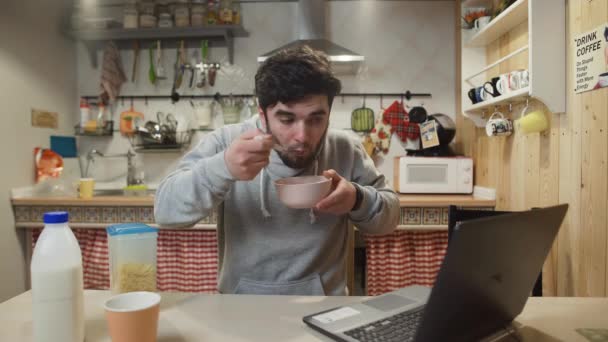 Hombre joven que trabaja ordenador portátil y come cereales de maíz en la cocina casera. — Vídeos de Stock