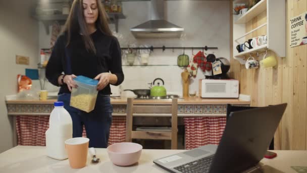 Mladá žena pracuje notebook a jí kukuřičné vločky obiloviny doma kuchyně. — Stock video