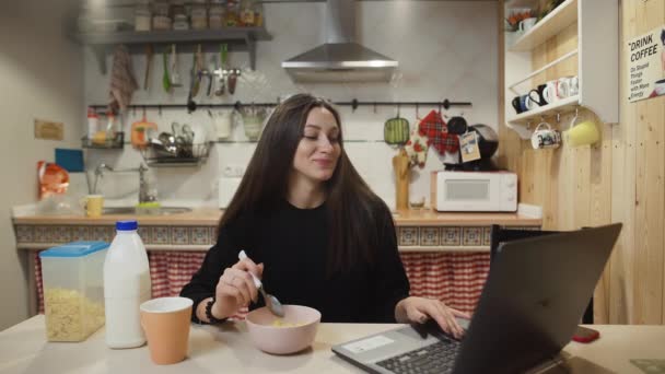 Mujer joven que trabaja ordenador portátil y come cereales de maíz en la cocina casera. — Vídeos de Stock