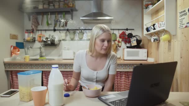 Ung kvinna tittar på video på bärbar dator och äter majsflingor spannmål hemma kök. — Stockvideo