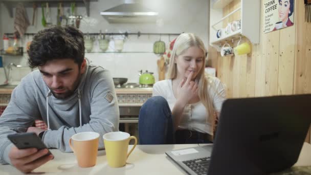 Fiatal pár beszél és használ modern laptop és telefon a konyhában. — Stock videók