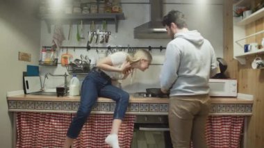 Genç koca tavada bir şeyler pişiriyor ve karısı ona gülüyor.